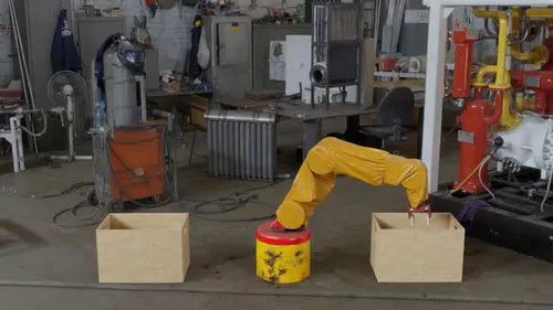 A multi-axes robot covered in yellow pvc fabric practising picking stuff up from one wooden box and placing it into another inside an industrial environment. The robot has a cylindrical base that is yellow with a red upper part starting around 80% up the cylinder. It is worn down with dark spots that look like dirt, rust, oil, or some combo of that. The top part of the cylinder has rounded edges. It doesn't actually pick anything up. 